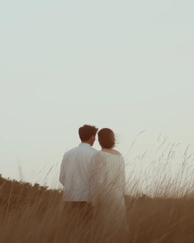 Couple standing together in a field at sunset, captured from behind in a soft, romantic portrait.