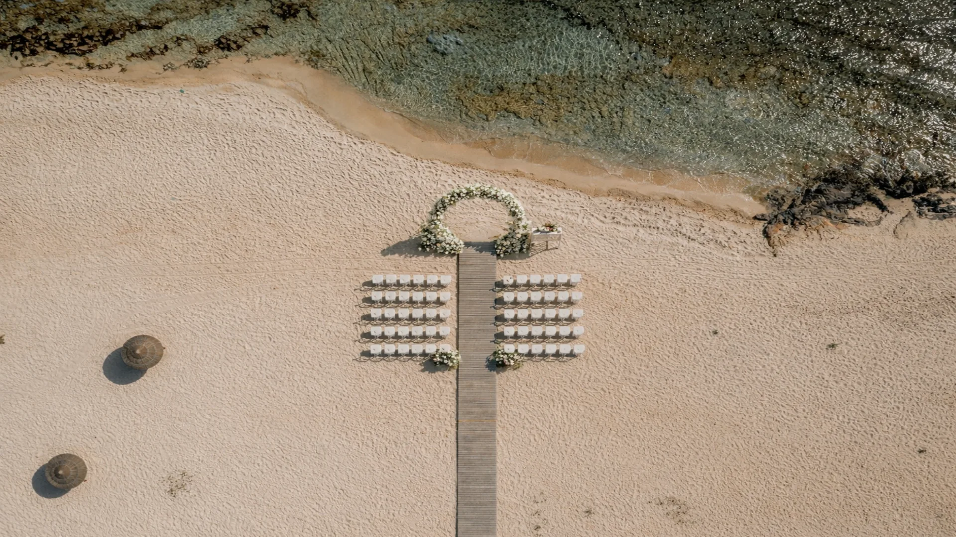 Aerial view of a beach wedding ceremony at Sirines Beach in Cyprus with circular floral arch, white chairs and wooden aisle