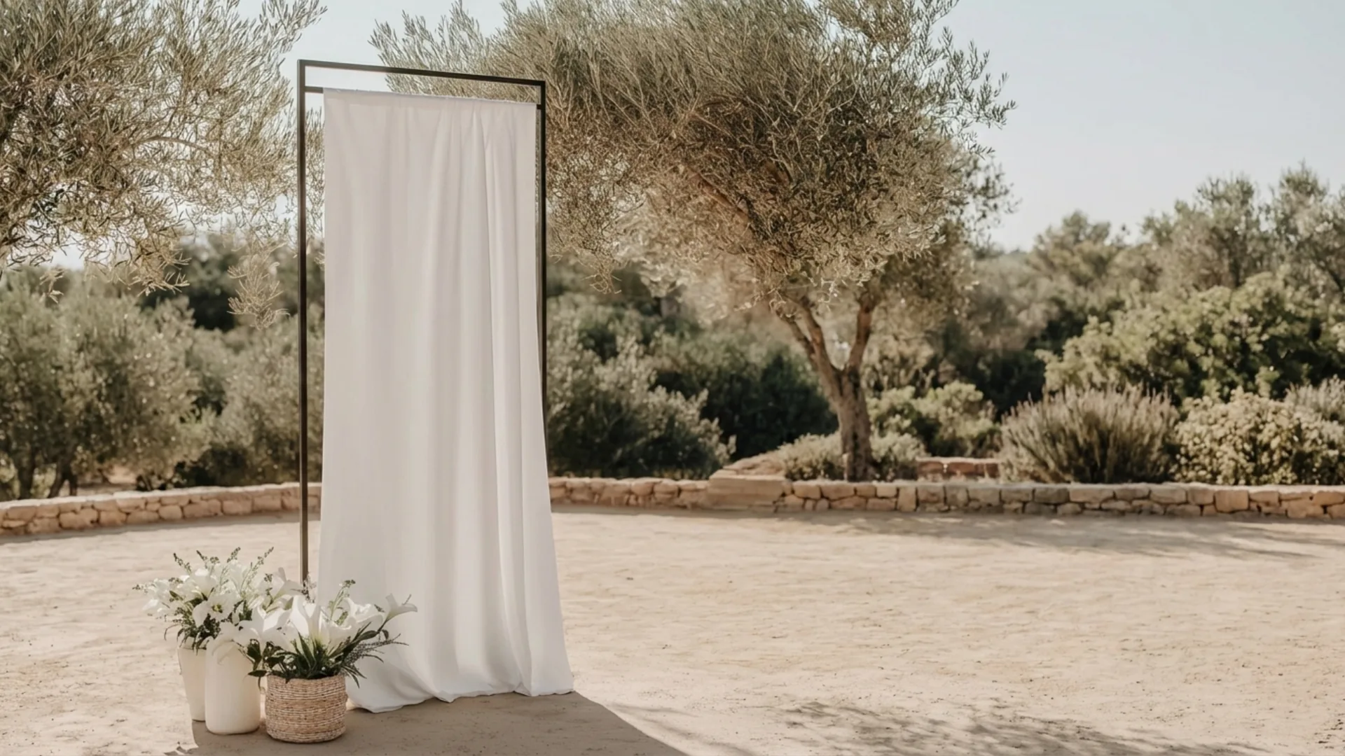 Minimalist outdoor wedding ceremony setup with white fabric backdrop and soft florals set among olive trees in a Mediterranean landscape by Soomore.