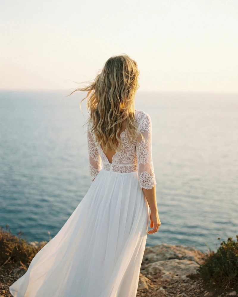 Bride in a flowing lace wedding dress standing by the Mediterranean Sea in Cyprus at golden hour, captured from behind in a serene coastal portrait.