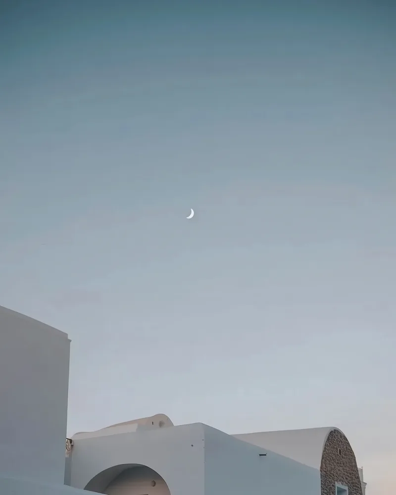 Minimal Mediterranean white architecture beneath a soft blue sky with a crescent moon, evoking calm evening atmosphere for a destination wedding setting.